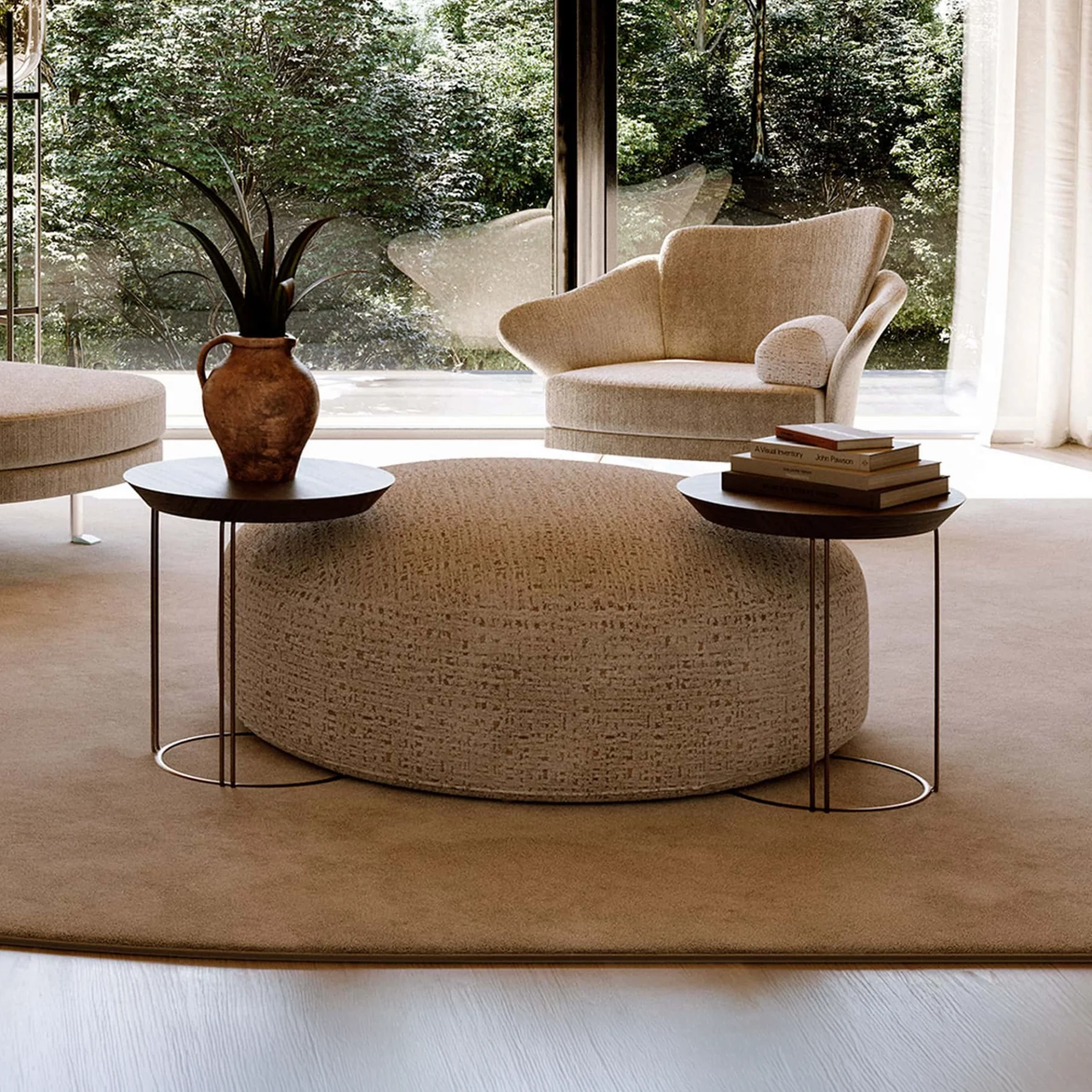 A modern living room with a large, round, textured beige ottoman, two dark wood side tables, and a sculptural armchair, all positioned in front of a floor-to-ceiling window overlooking lush green trees.