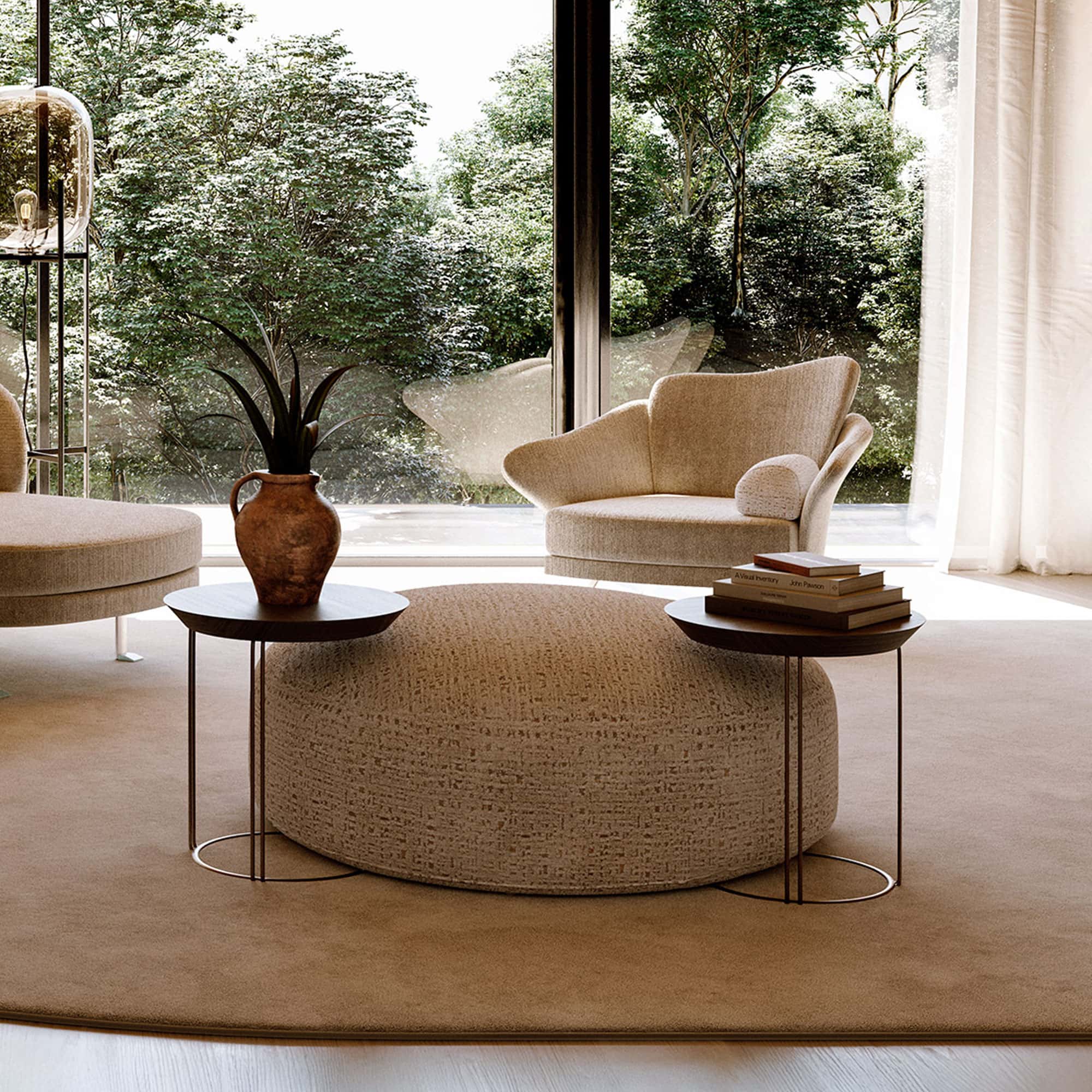 A modern living room with a large, round, textured beige ottoman, two dark wood side tables, and a sculptural armchair, all positioned in front of a floor-to-ceiling window overlooking lush green trees.