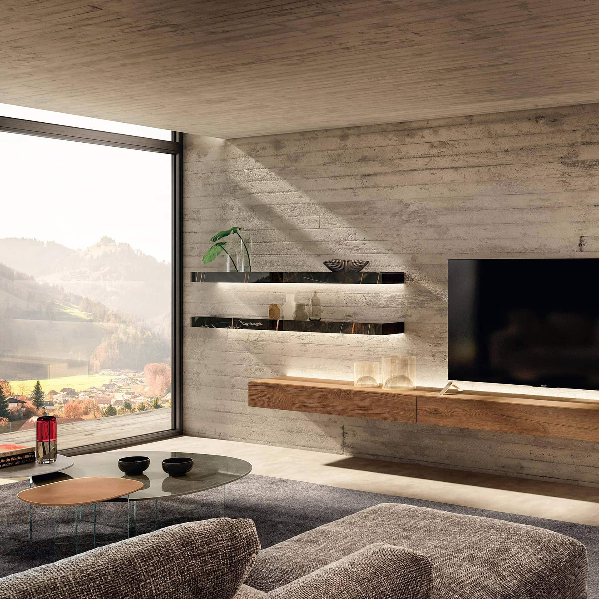 A modern living room with concrete walls and ceiling, featuring a large window overlooking a scenic valley. The room has floating shelves with LED lighting, a wooden media console with a TV, and a sofa with a coffee table on a grey rug.