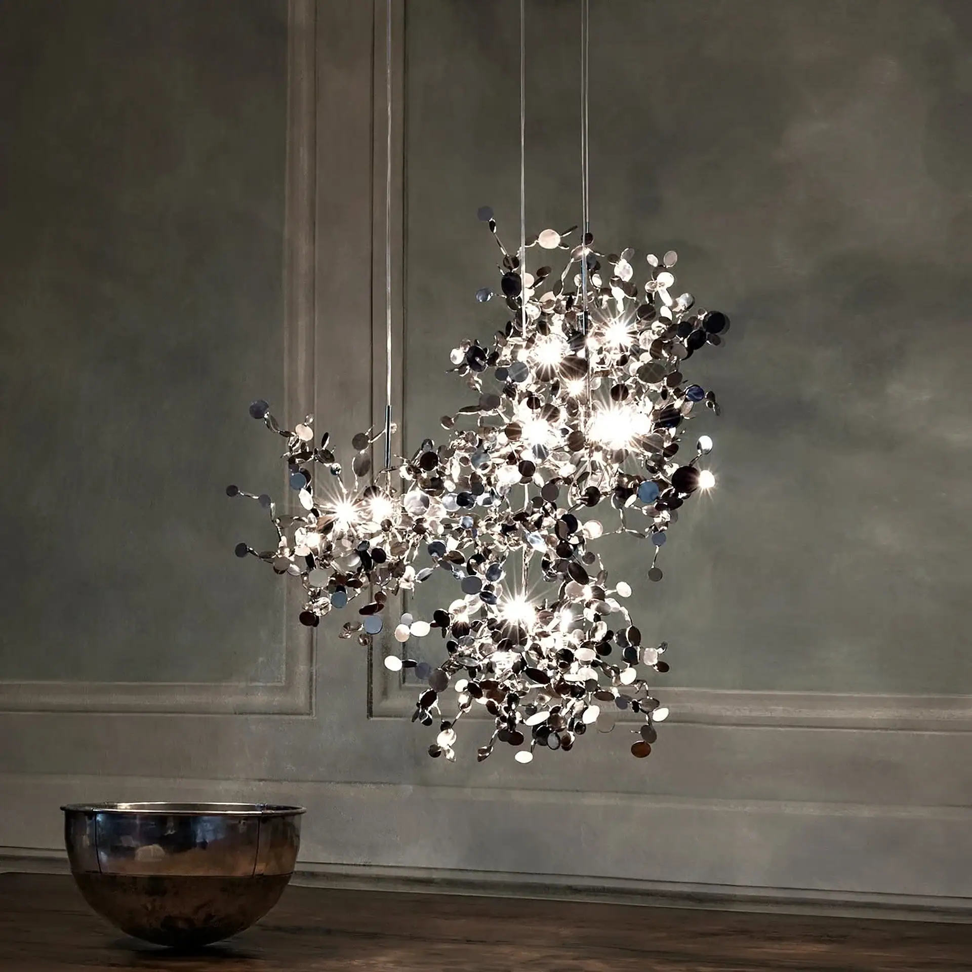 A modern chandelier with numerous metallic discs and bright lights hangs in a room with dark walls and a wooden floor. A metallic bowl sits on the floor.