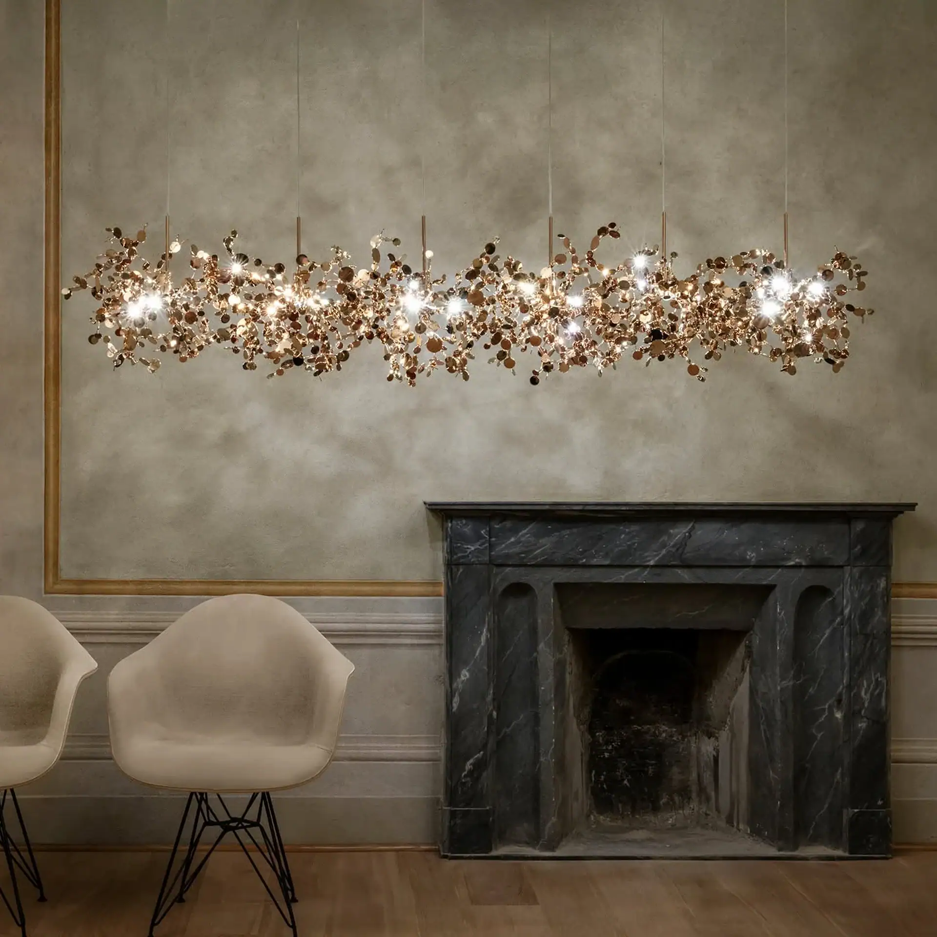 A long, horizontal chandelier made of many small, shiny copper discs and lights hangs above a dark gray marble fireplace. Two light beige chairs are in the foreground.
