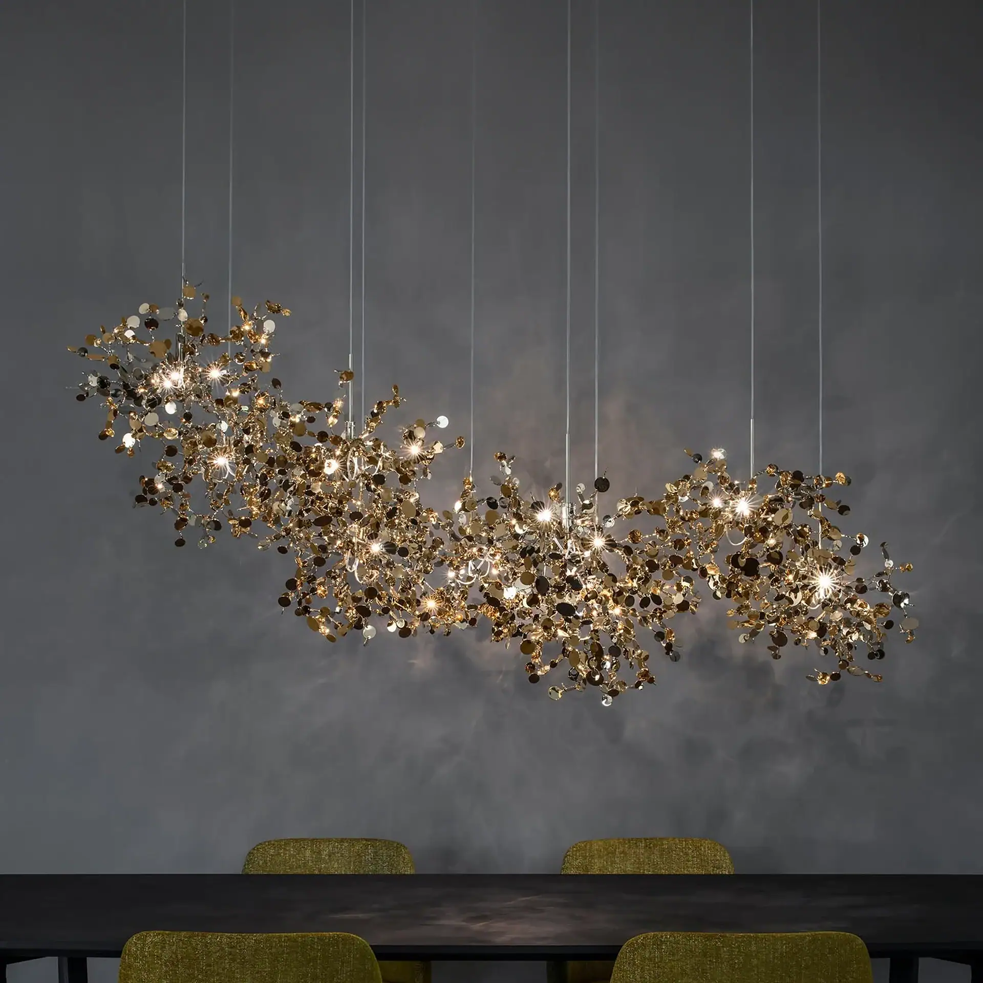 A large, organic-shaped chandelier with gold and silver reflective discs and multiple lights hangs over a dark table and two green chairs against a dark gray wall.
