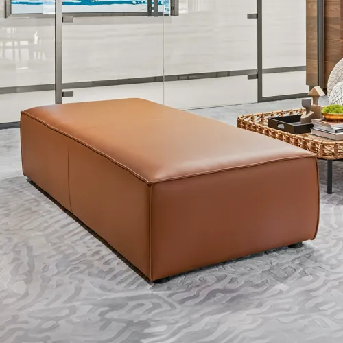 A long, rectangular brown leather ottoman on a gray patterned rug in a modern room with a glass partition and a wicker coffee table.