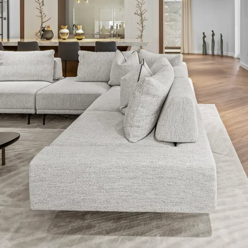 A large, light gray textured sectional sofa in a modern living room. The sofa sits on a light rug, with hardwood flooring and a kitchen counter visible in the background.