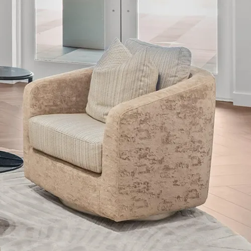 A contemporary, rounded swivel armchair upholstered in textured beige fabric with lighter patterned cushions, sitting on a light rug in a bright room.