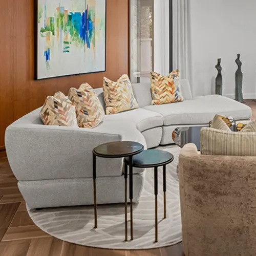 A modern living room with a light gray curved sectional sofa, patterned throw pillows, an abstract painting on a wooden wall, and two round nesting side tables.