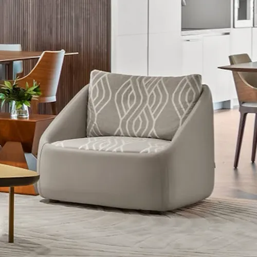 A modern, armless gray accent chair with patterned cushions sits on a light rug in a contemporary living room, with dining furniture and a kitchen visible in the background.