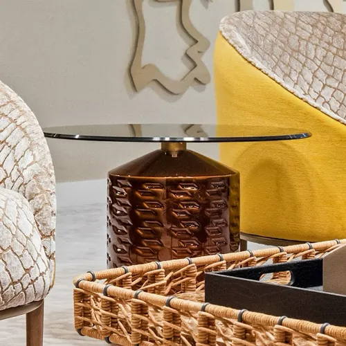A close-up of a modern interior with a glass-top side table featuring a textured dark brown base, flanked by a yellow armchair and a neutral-toned chair, with a woven tray in the foreground.
