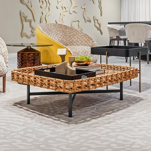 A modern living room with a square woven rattan coffee table, a yellow and patterned armchair, and abstract wall art. The table holds a black tray with decorative sculptures and a moss bowl.