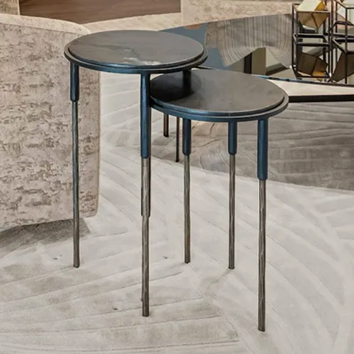 Two modern, dark round nesting side tables with slender two-toned metal legs, placed on a light-colored patterned rug in a contemporary living space.