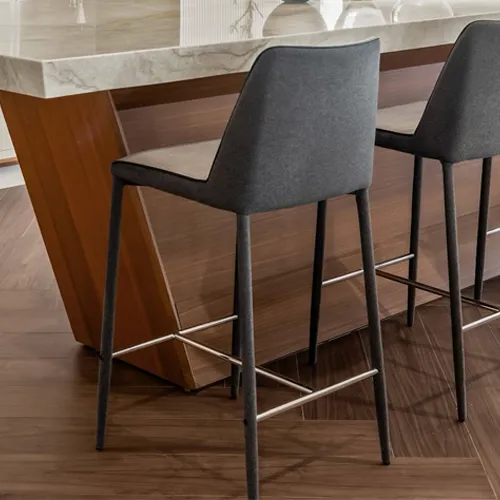 Two dark gray upholstered bar stools with metal legs and footrests are positioned at a modern kitchen island. The island features a warm-toned, angular wood base and a light marble countertop. The flooring is dark brown wood.