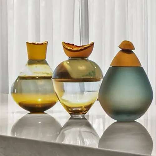 Three decorative glass vessels with amber, green, and teal colors on a reflective surface, against a blurred white curtain background.