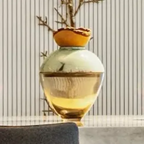 Three decorative glass vessels with amber, green, and teal colors on a reflective surface, against a blurred white curtain background.