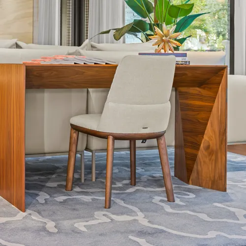 A modern wooden console table with an angular leg design, paired with a light beige upholstered chair, on a grey abstract patterned rug. Green plants and a window are in the background.