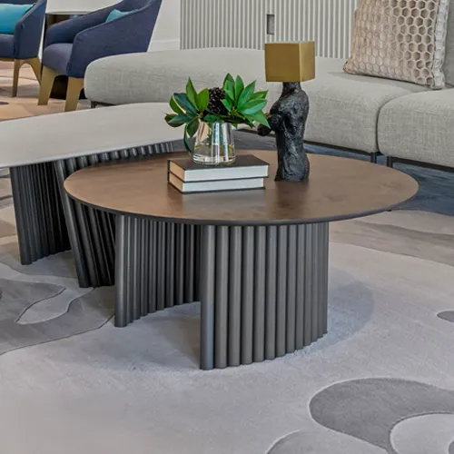 A round coffee table with a dark bronze top and a base made of vertical gray rods. It holds a vase with greenery, stacked books, and an abstract sculpture. A gray sofa, bench, and patterned rug are visible in the background.