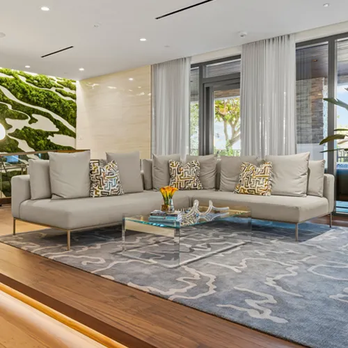 A modern living room with a light grey L-shaped sofa, glass coffee table, blue-grey patterned rug, and a large green moss wall art installation on the left.