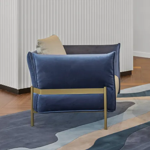 A luxurious deep blue velvet armchair with gold metal legs sits on an abstract blue and beige rug in a modern room with white fluted walls.