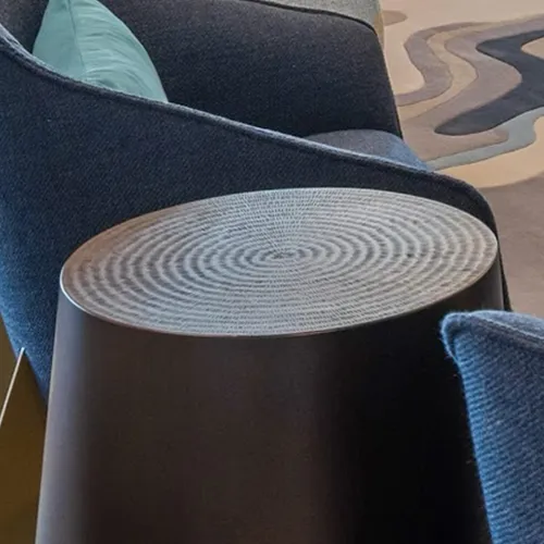 A dark cylindrical side table with a light gray concentric circular pattern on its top, positioned next to a dark blue sofa with a teal throw pillow. An abstract patterned rug is visible in the background.