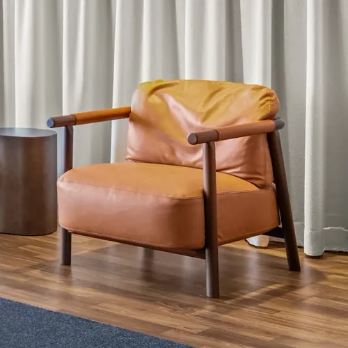 A modern armchair with cognac leather cushions and a dark wooden frame, next to a cylindrical side table, on a hardwood floor with a partial rug, in front of light curtains.