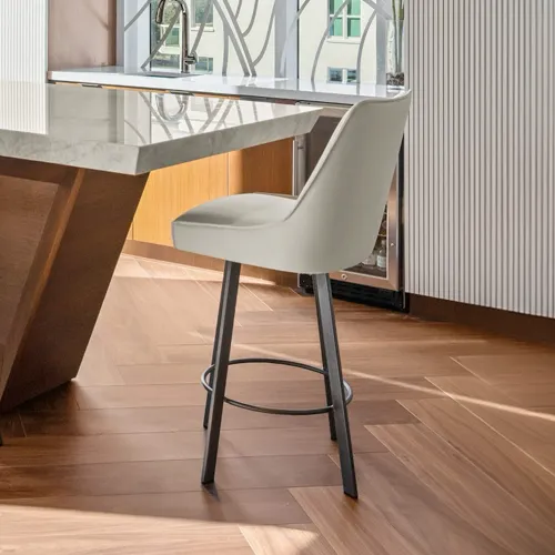 A modern kitchen scene featuring a light grey upholstered bar stool next to a marble-topped kitchen island with a wooden base and herringbone flooring.