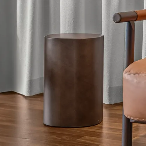 A dark brown cylindrical side table stands on a wooden floor, flanked by light gray curtains and a tan leather armchair.