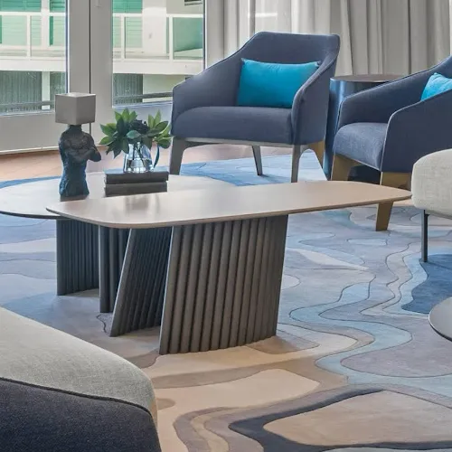 A modern lounge with a light-colored coffee table featuring a ribbed base, surrounded by dark blue armchairs with turquoise pillows, and an abstract blue and gray rug.