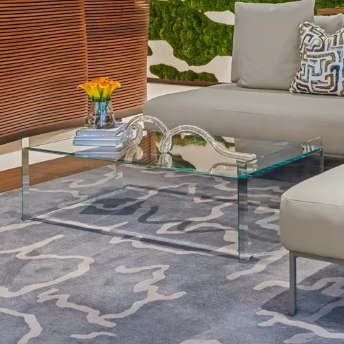 A modern living room with a glass coffee table, orange flowers, a light sofa with a patterned pillow, and a green moss wall feature.