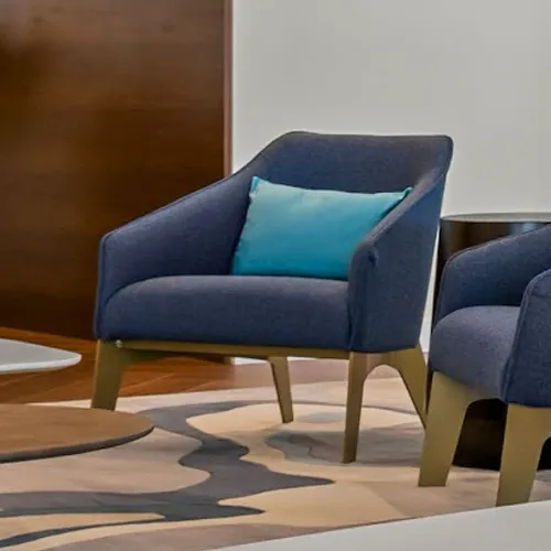 A navy blue upholstered armchair with a bright teal pillow, set on an abstract patterned rug in a modern lounge area with wooden accents.