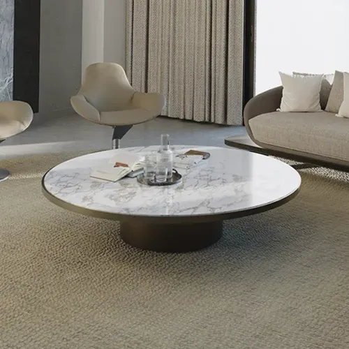 A modern living room with a round white marble coffee table, a beige armchair, and a light-colored sofa on a textured rug. A glass carafe and book are on the table.