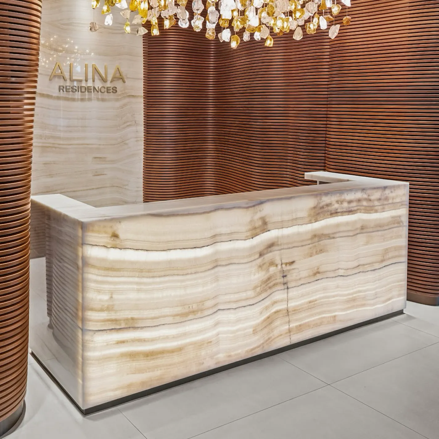 A luxurious modern lobby featuring a glowing translucent onyx reception desk, warm horizontal wooden slat walls, and a sparkling chandelier made of clear and amber crystal pieces. 'ALINA RESIDENCES' is visible in gold lettering on the back wall.