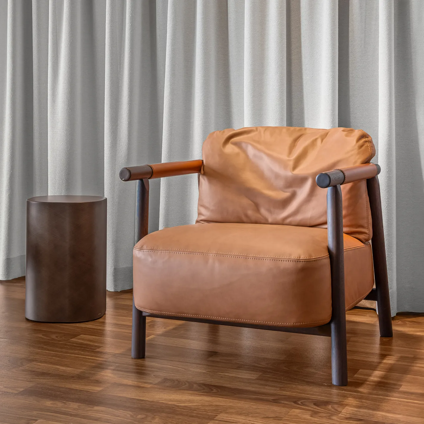 A modern tan leather armchair with a dark wood frame next to a dark cylindrical side table, set against light grey pleated curtains and a warm wooden floor.