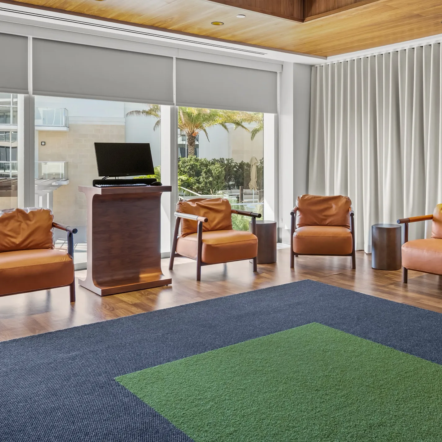 A modern lounge with four caramel leather armchairs, a wooden podium with a monitor, a blue and green geometric rug, and large windows overlooking palm trees and white buildings.