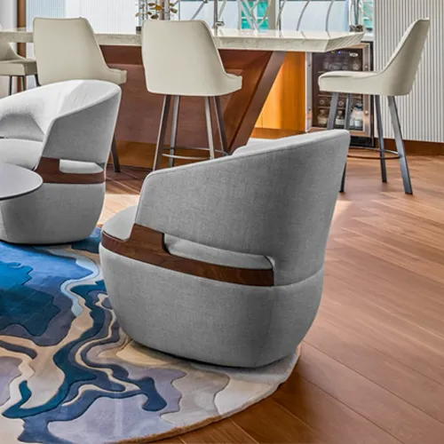 A modern lounge area with two gray upholstered swivel chairs featuring wood accents, placed on a round blue and white abstract rug. In the background, a bar with a light countertop and cream bar stools is visible.