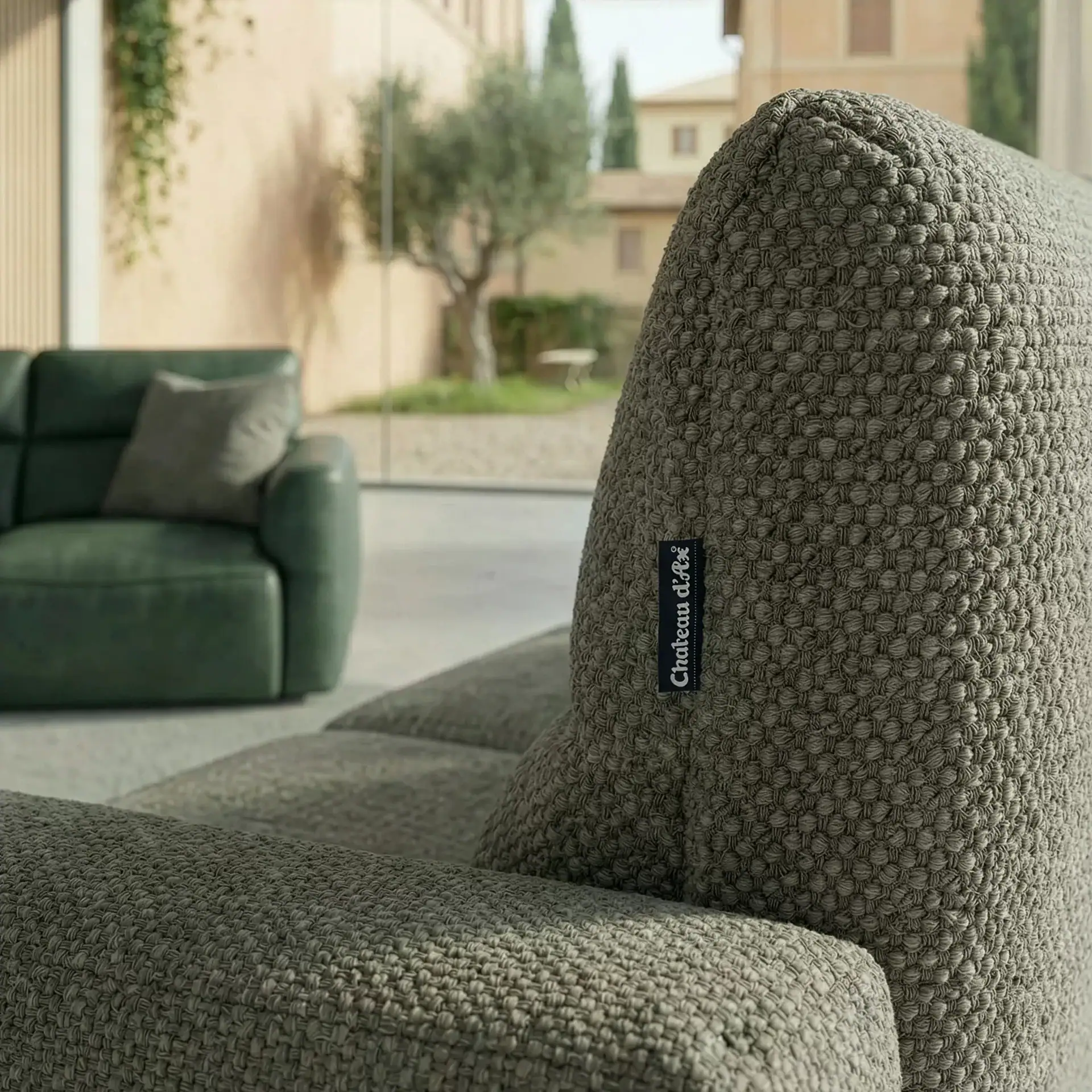 Close-up of a grey-green textured sofa with a 'Chateau d'Ax' label, a blurred green sofa, and an outdoor view in the background.