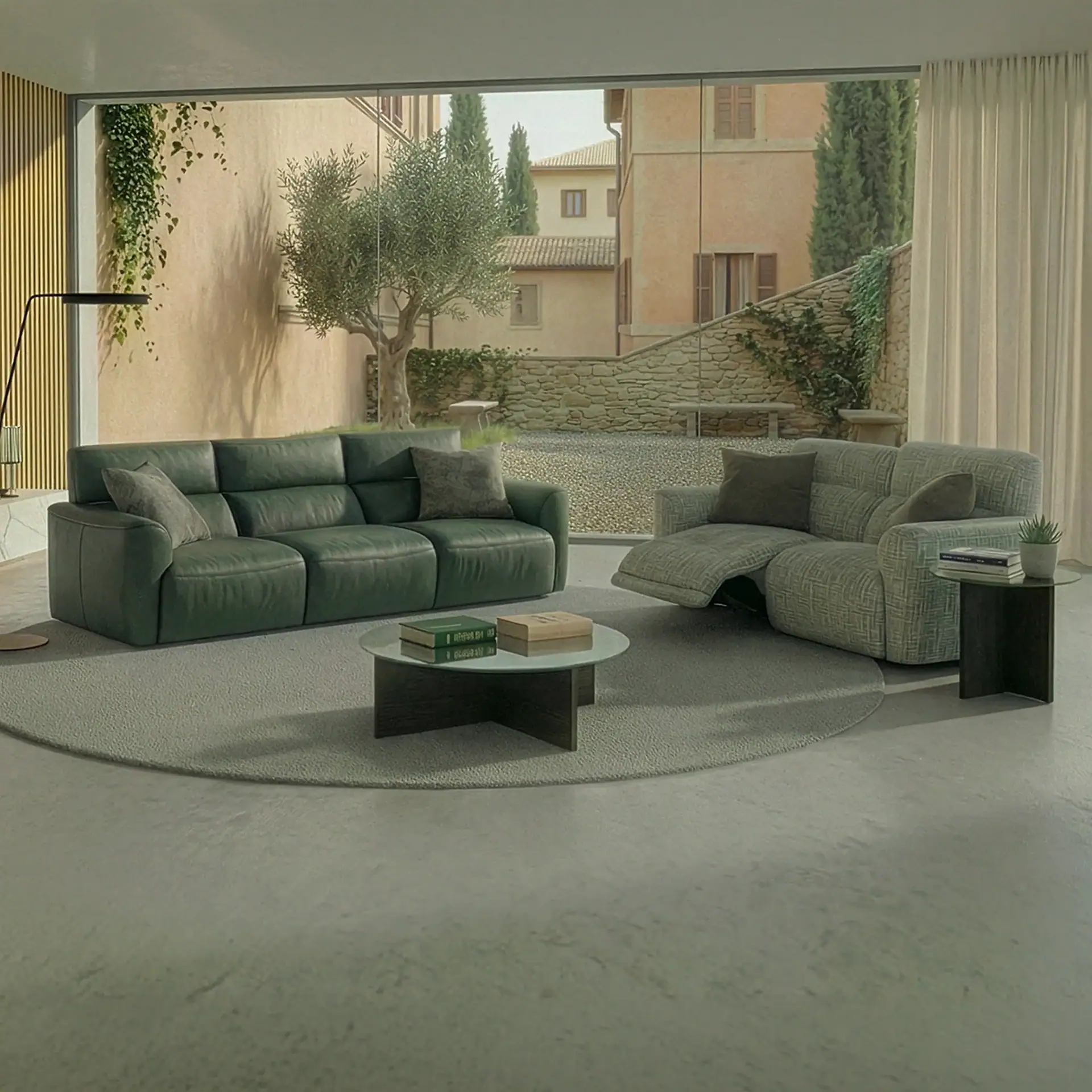 A modern living room with a green leather sofa, a gray recliner, and a round coffee table on a rug, overlooking an outdoor courtyard with olive trees and buildings through a large window.