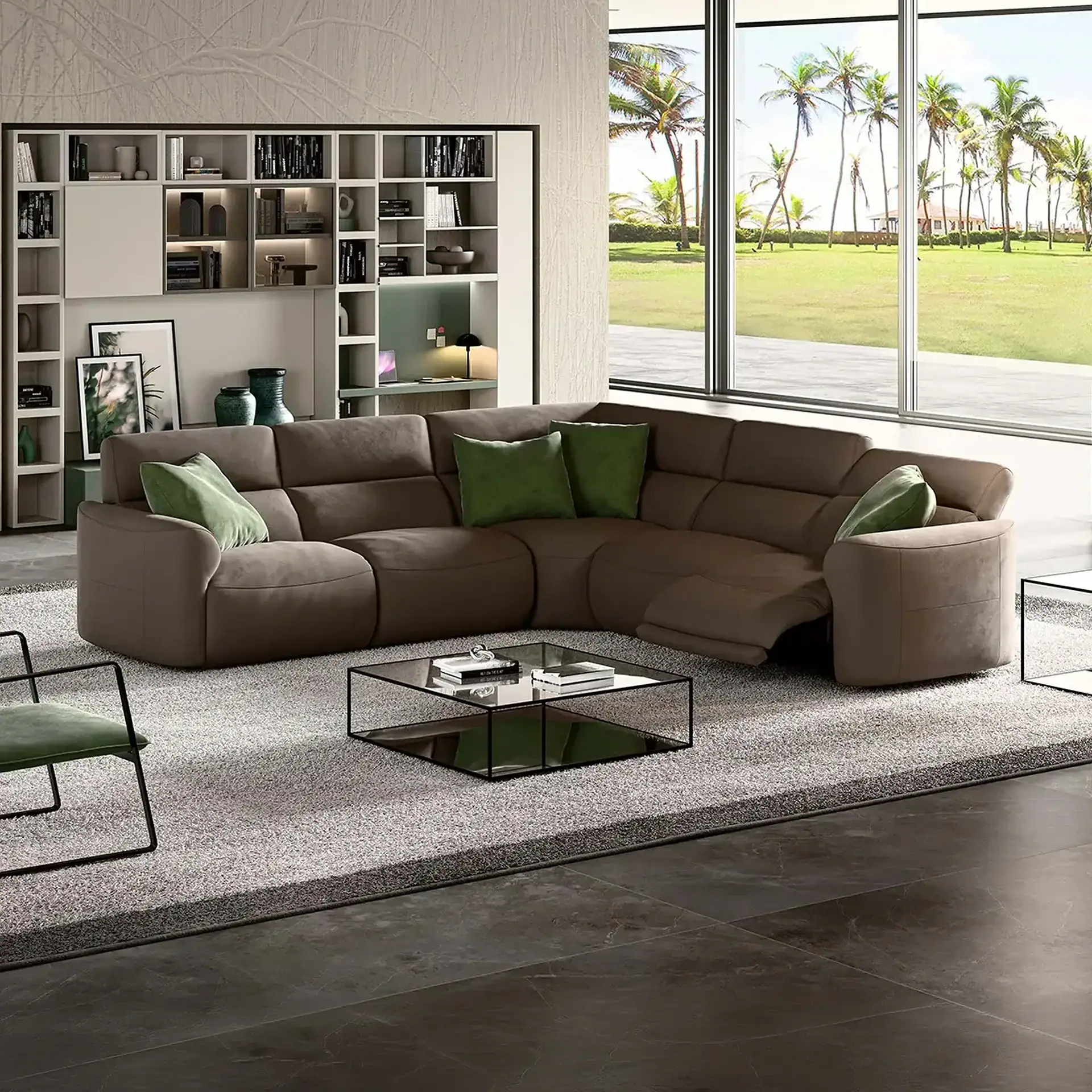 A modern living room with a large brown L-shaped sectional sofa, green pillows, a glass coffee table, and a built-in shelving unit. Large windows overlook a tropical landscape with palm trees and a green lawn.