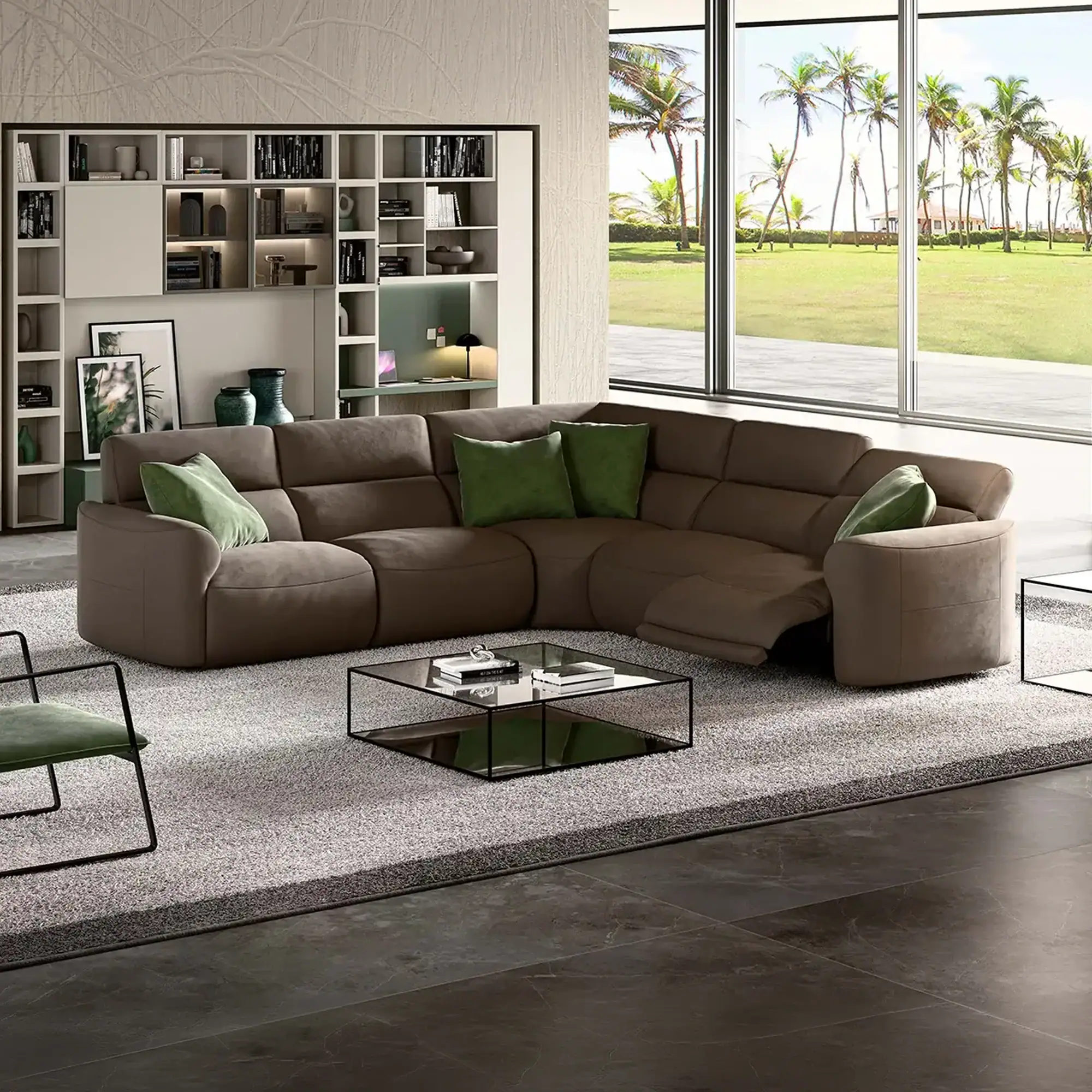 A modern living room with a large brown L-shaped sectional sofa, green pillows, a glass coffee table, and a built-in shelving unit. Large windows overlook a tropical landscape with palm trees and a green lawn.