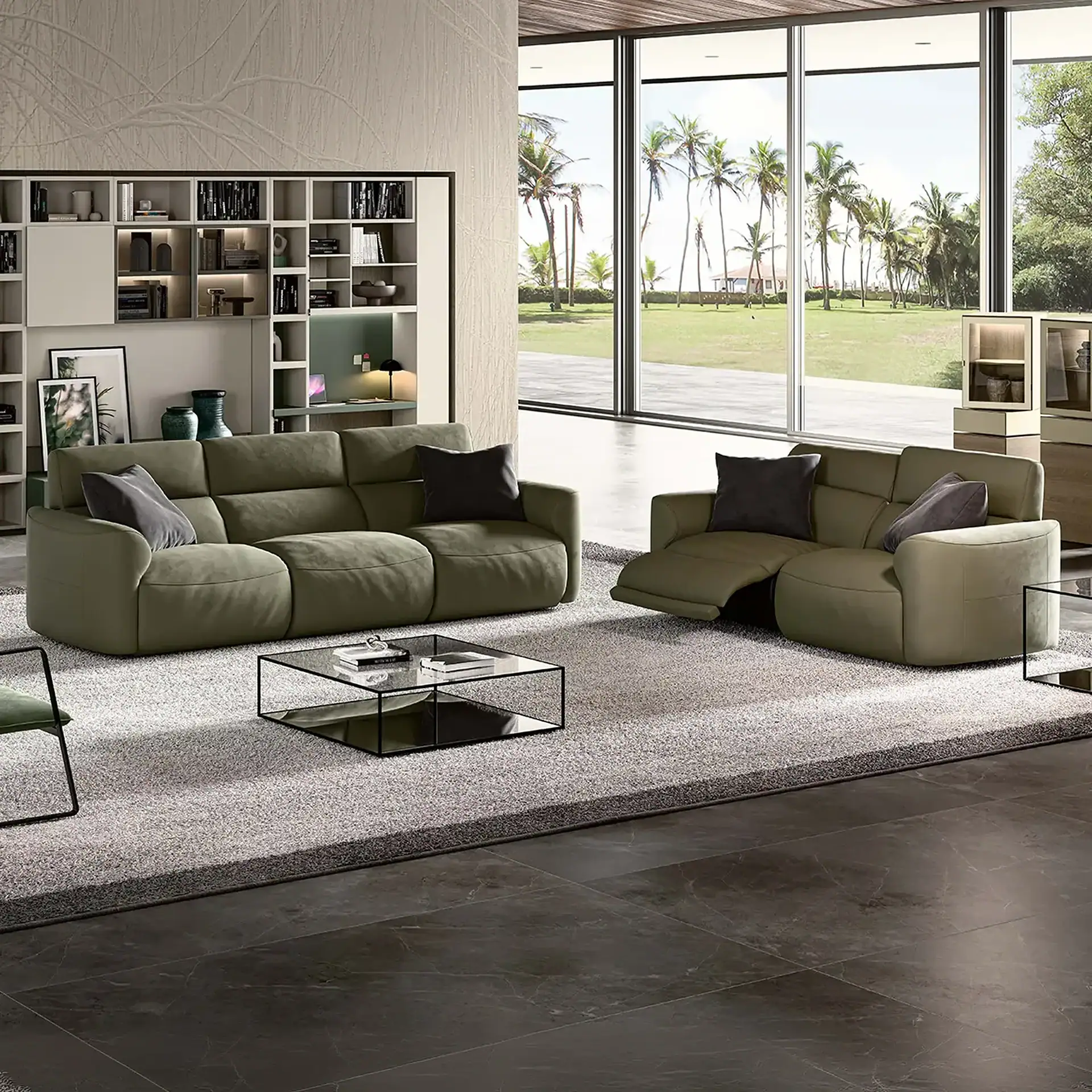 A modern living room with two olive green reclining sofas, a glass coffee table, and a large window overlooking a lawn with palm trees.