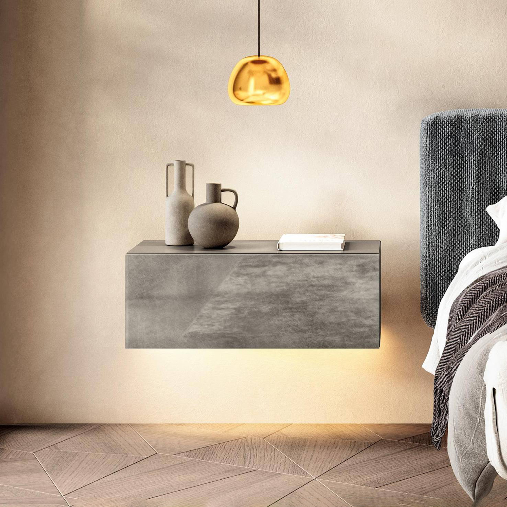 A modern bedroom scene featuring a floating gray nightstand with underlighting, a golden pendant lamp, and a bed with a textured headboard and patterned throw. Two ceramic vases and a book adorn the nightstand.