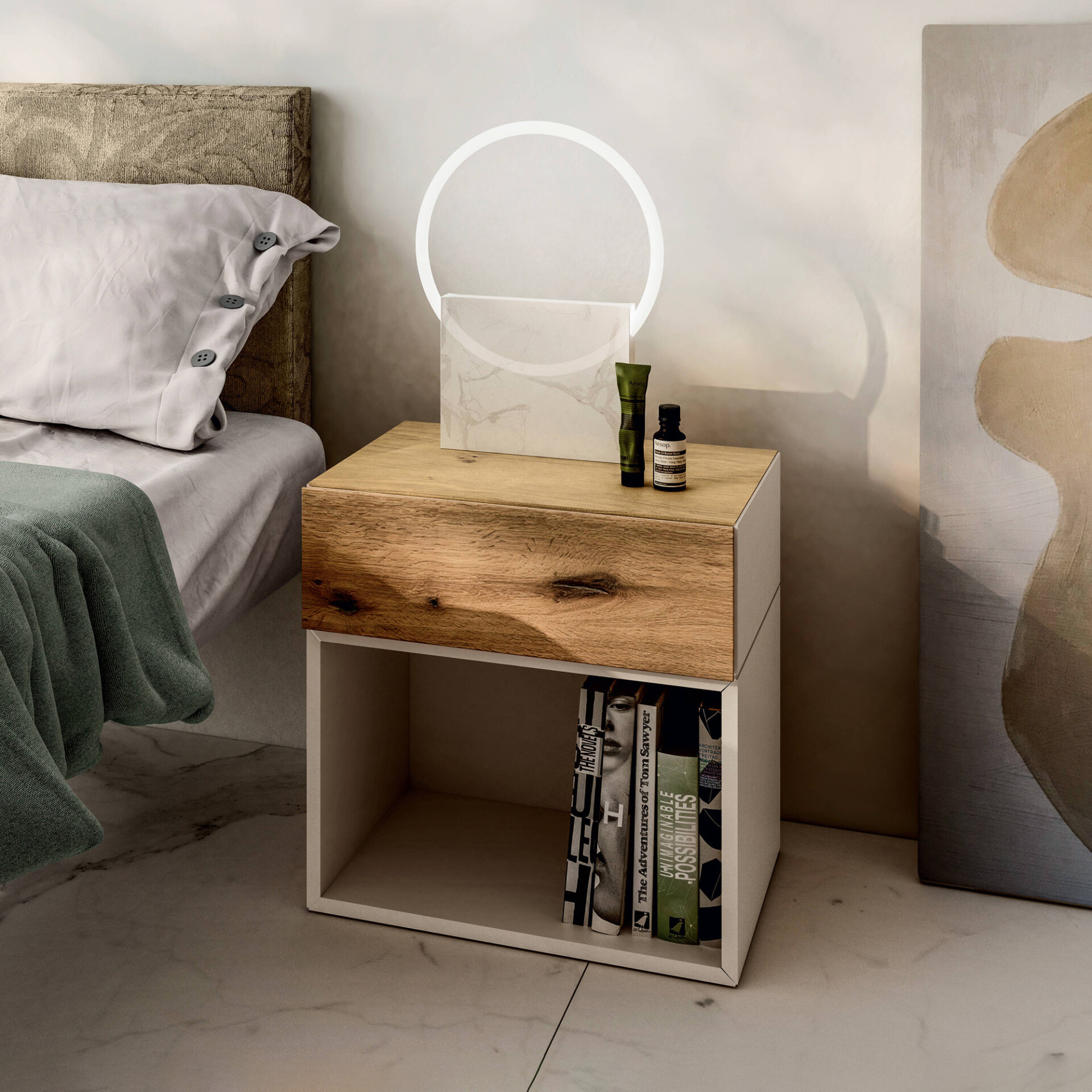 A modern bedroom scene featuring a wooden nightstand with a circular lamp and Aesop products, next to a bed with a grey pillow and green throw blanket. An abstract artwork leans against the wall.