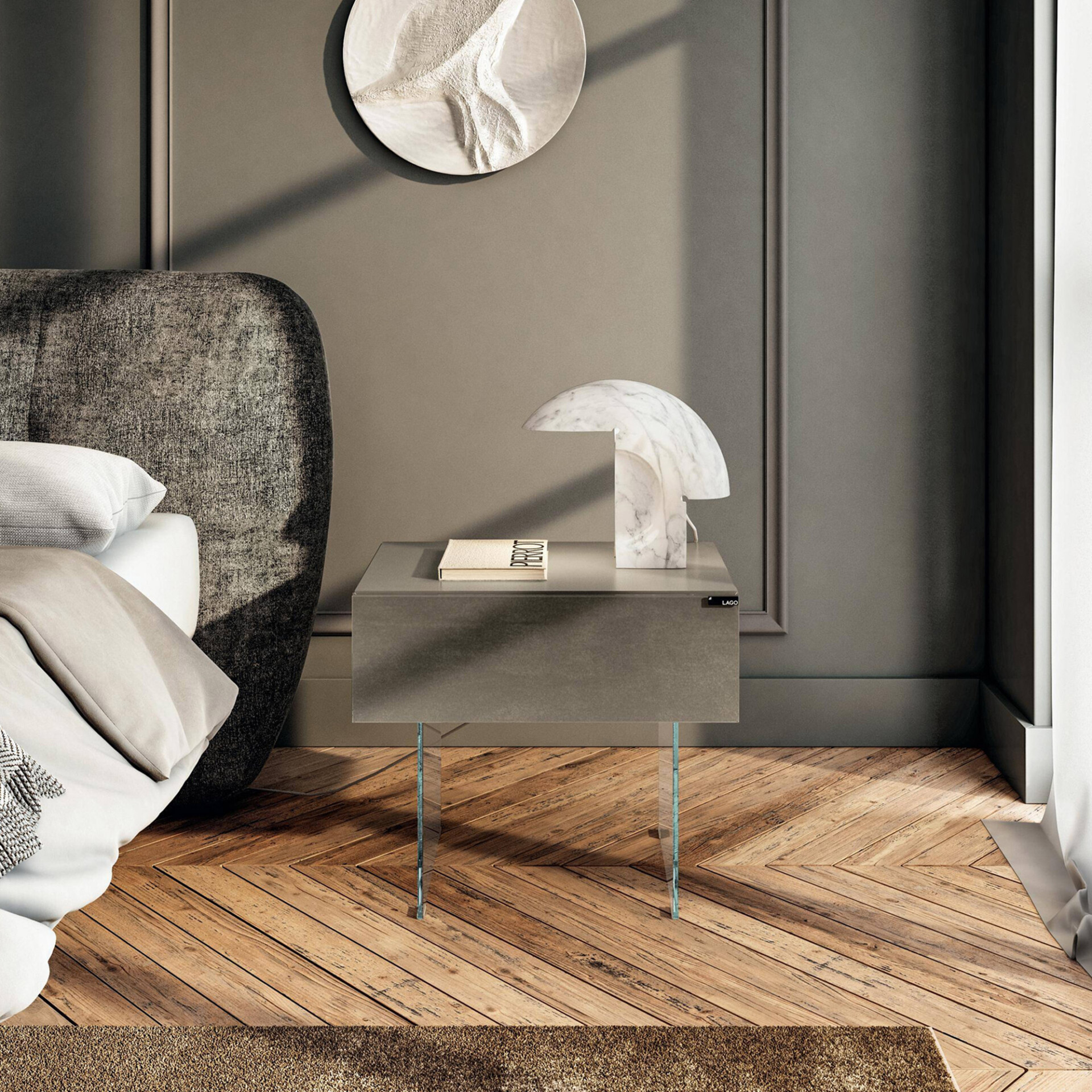 A modern bedroom with a dark grey upholstered bed, a minimalist dark grey nightstand with clear glass legs, and a white marble mushroom lamp. A textured white circular art piece hangs on the taupe paneled wall, above a herringbone wooden floor.