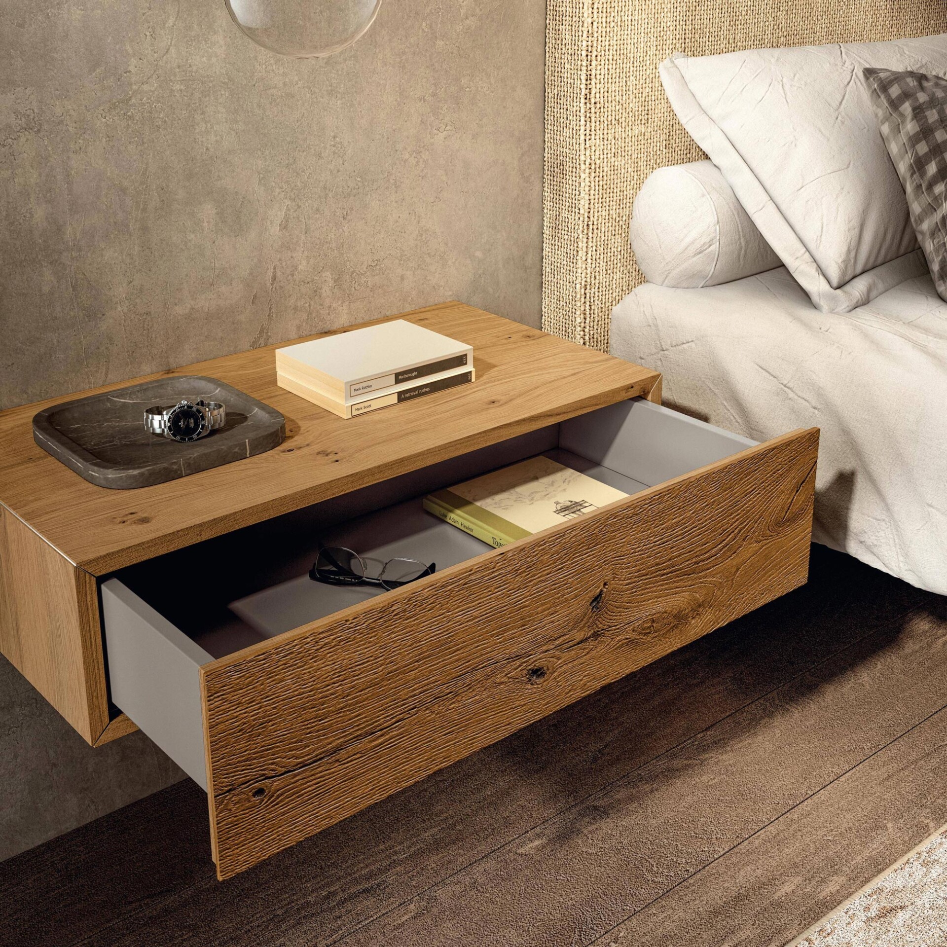 A floating wooden nightstand with an open drawer next to a bed with a woven headboard. On the nightstand are a watch on a tray and stacked books; eyeglasses and a book are inside the drawer.