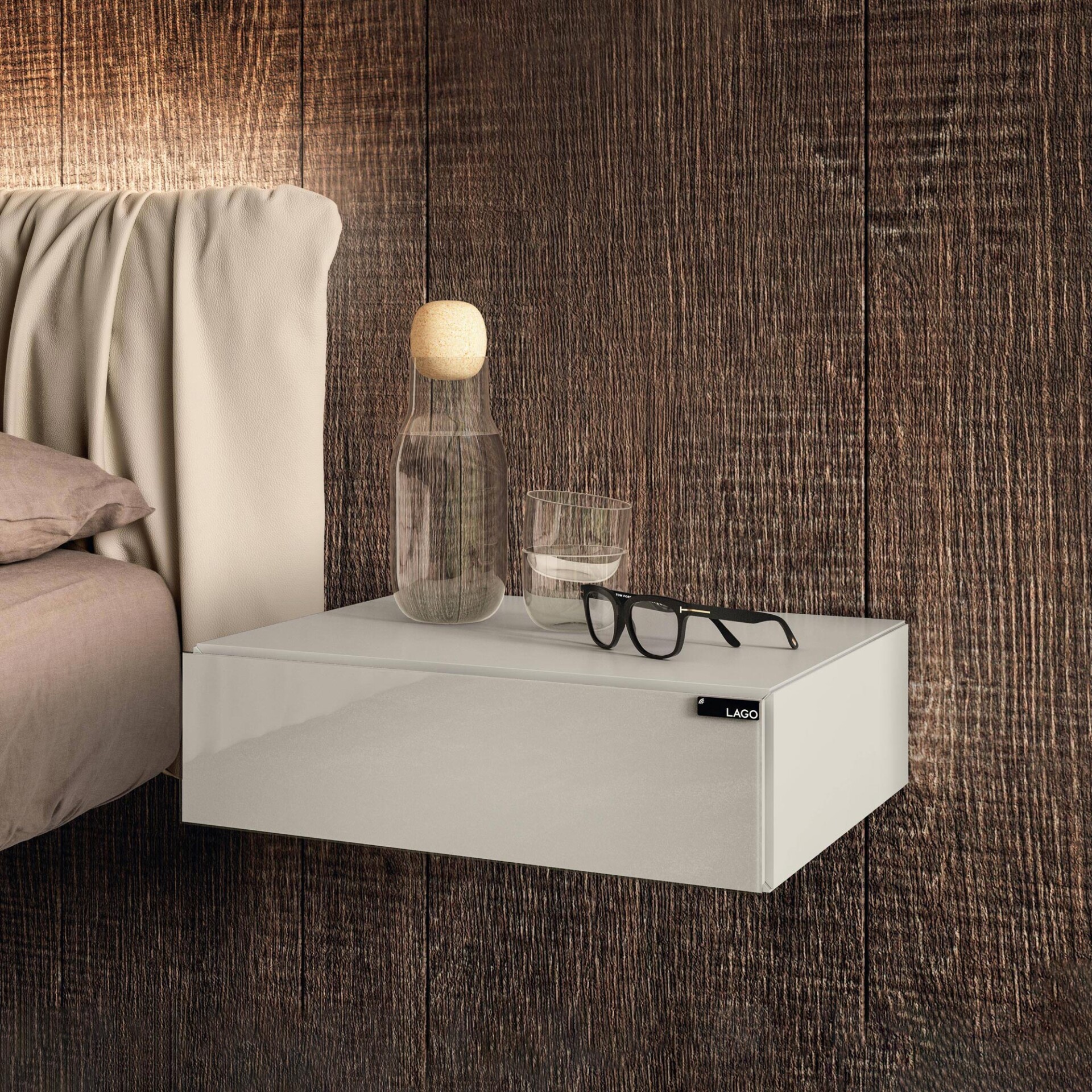 A modern floating light greige bedside table with a LAGO logo, holding a water bottle, a glass of water, and black eyeglasses, set against a dark wooden wall and a light upholstered headboard.