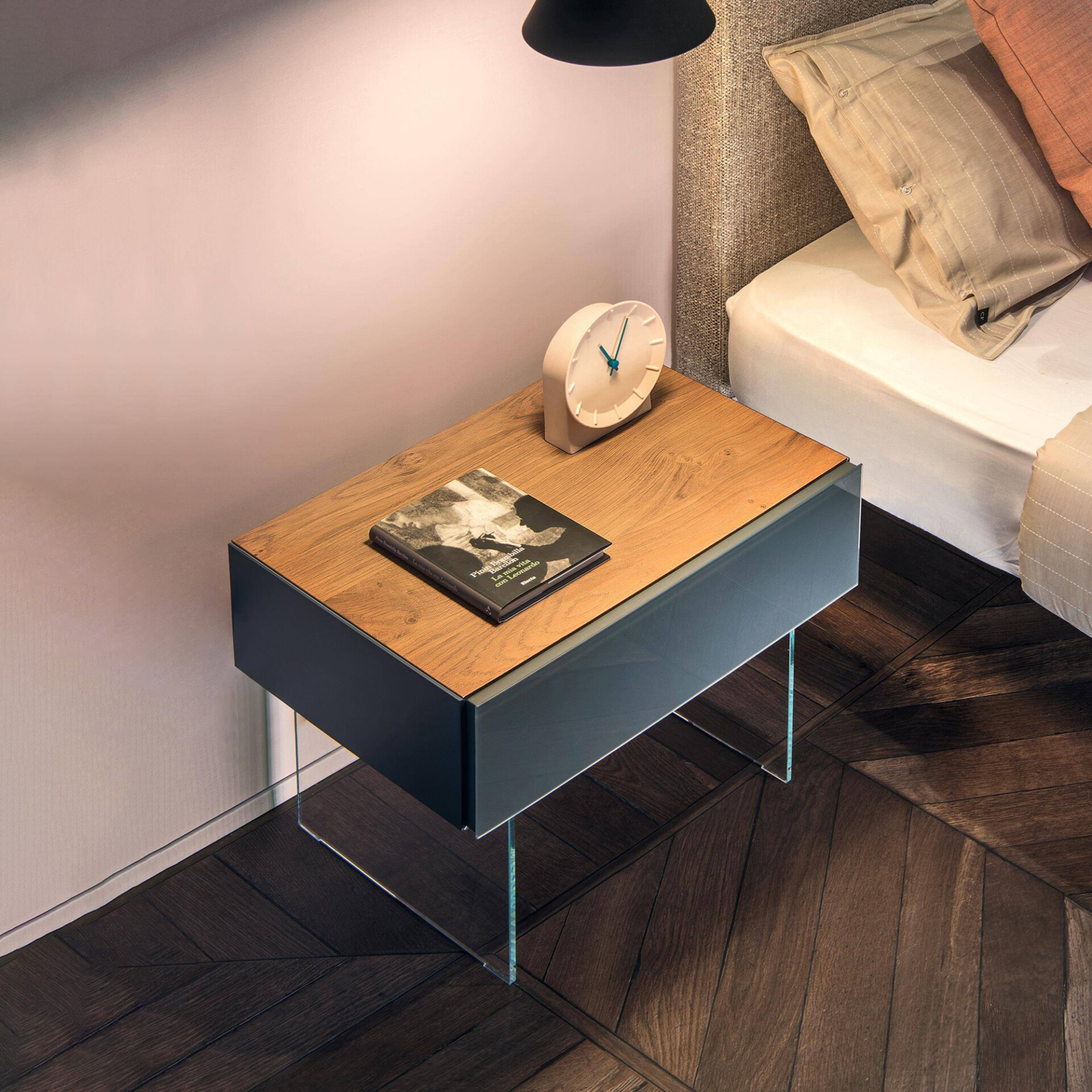 A modern bedroom scene with a floating nightstand featuring a wooden top, dark body, and glass legs. A book and a white clock sit on the nightstand, next to a bed with textured pillows, all illuminated by a black pendant lamp. The floor is dark herringbone wood.