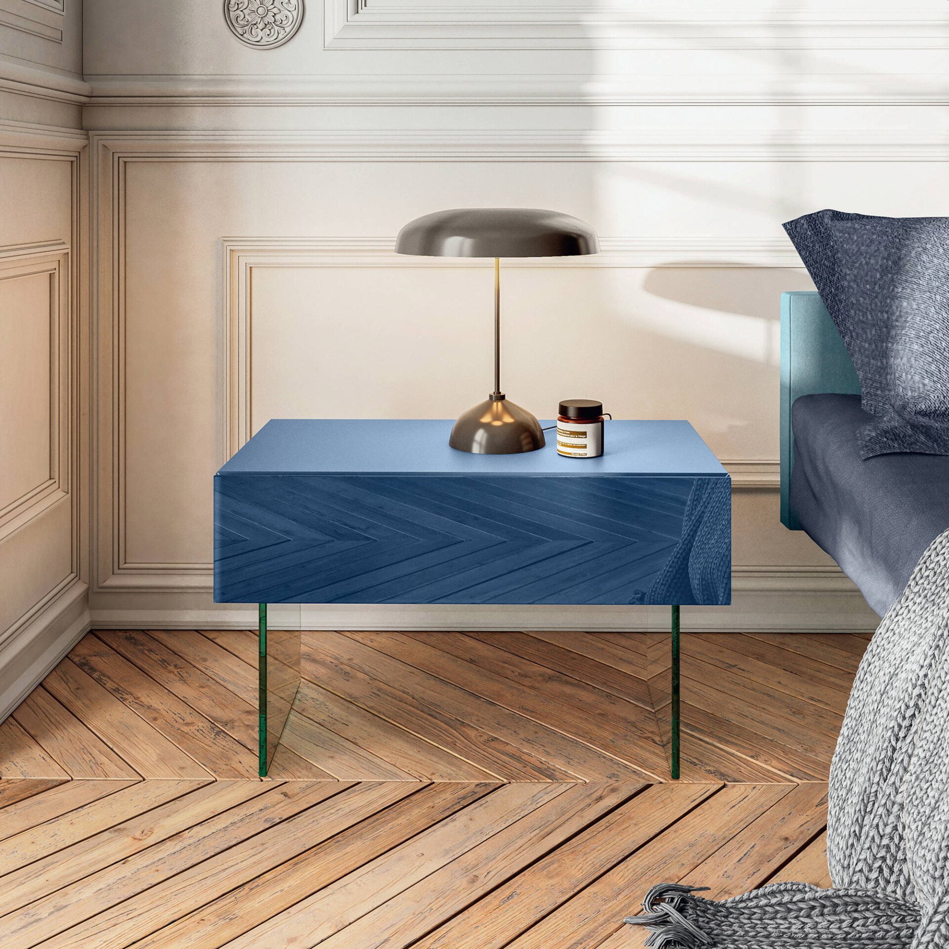 A modern deep blue nightstand with a chevron pattern and clear glass legs, topped with a dark mushroom-shaped lamp and a small jar. It sits in a bedroom with classic white paneled walls, a partial view of a bed with blue bedding, and a light wood herringbone floor.