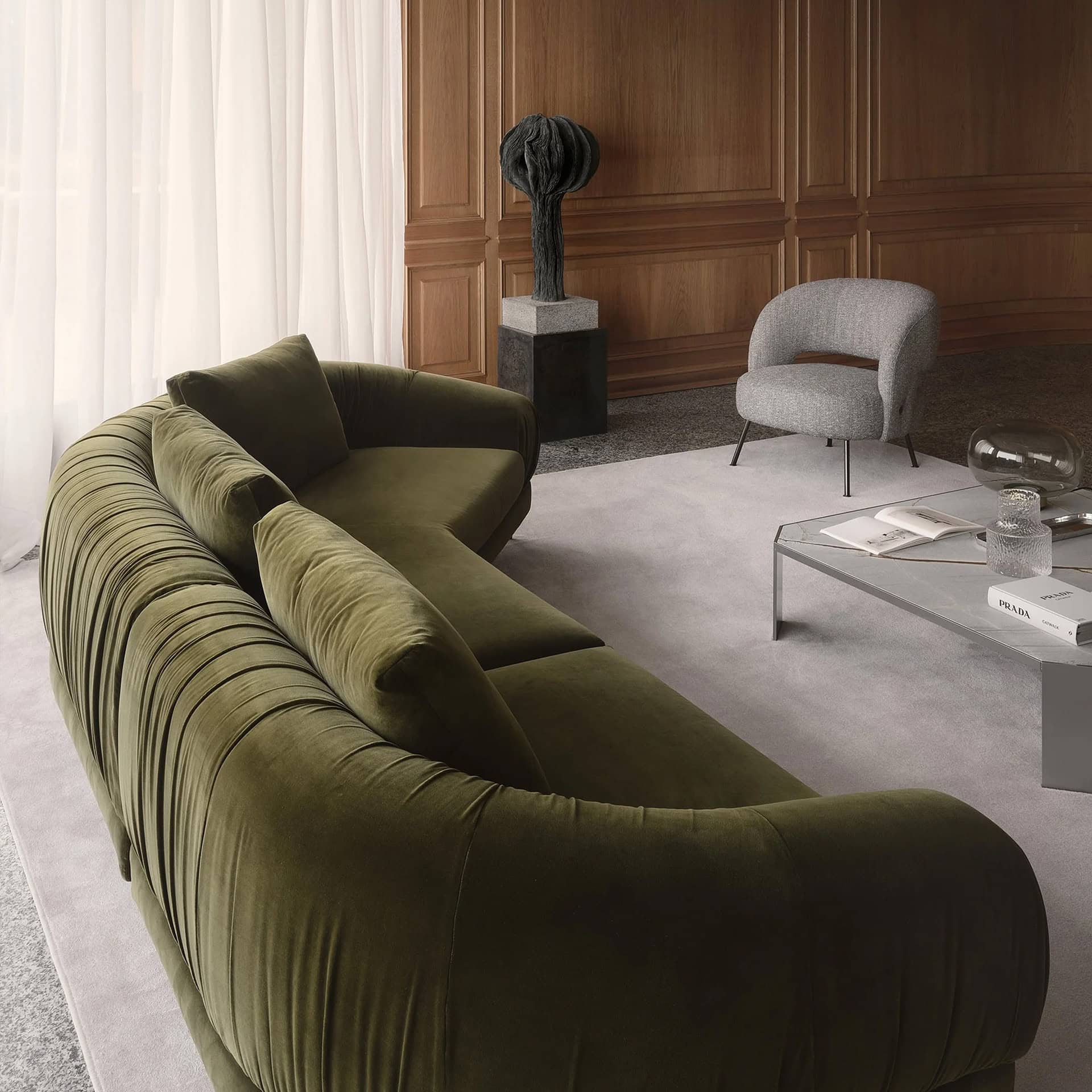 A modern living room featuring a large, curved olive green velvet sofa, a light grey coffee table with books and decor, a grey armchair, and a dark abstract sculpture against a warm wooden paneled wall. Sheer white curtains are visible on the left.