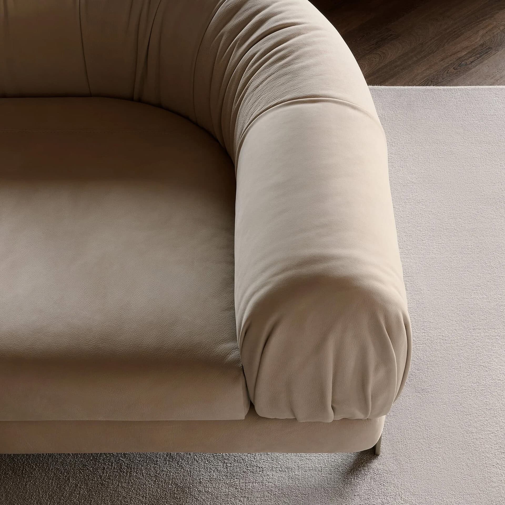 Close-up of a plush, light beige sofa armrest with soft, rounded curves, resting on a light-colored carpet next to a dark wooden floor.