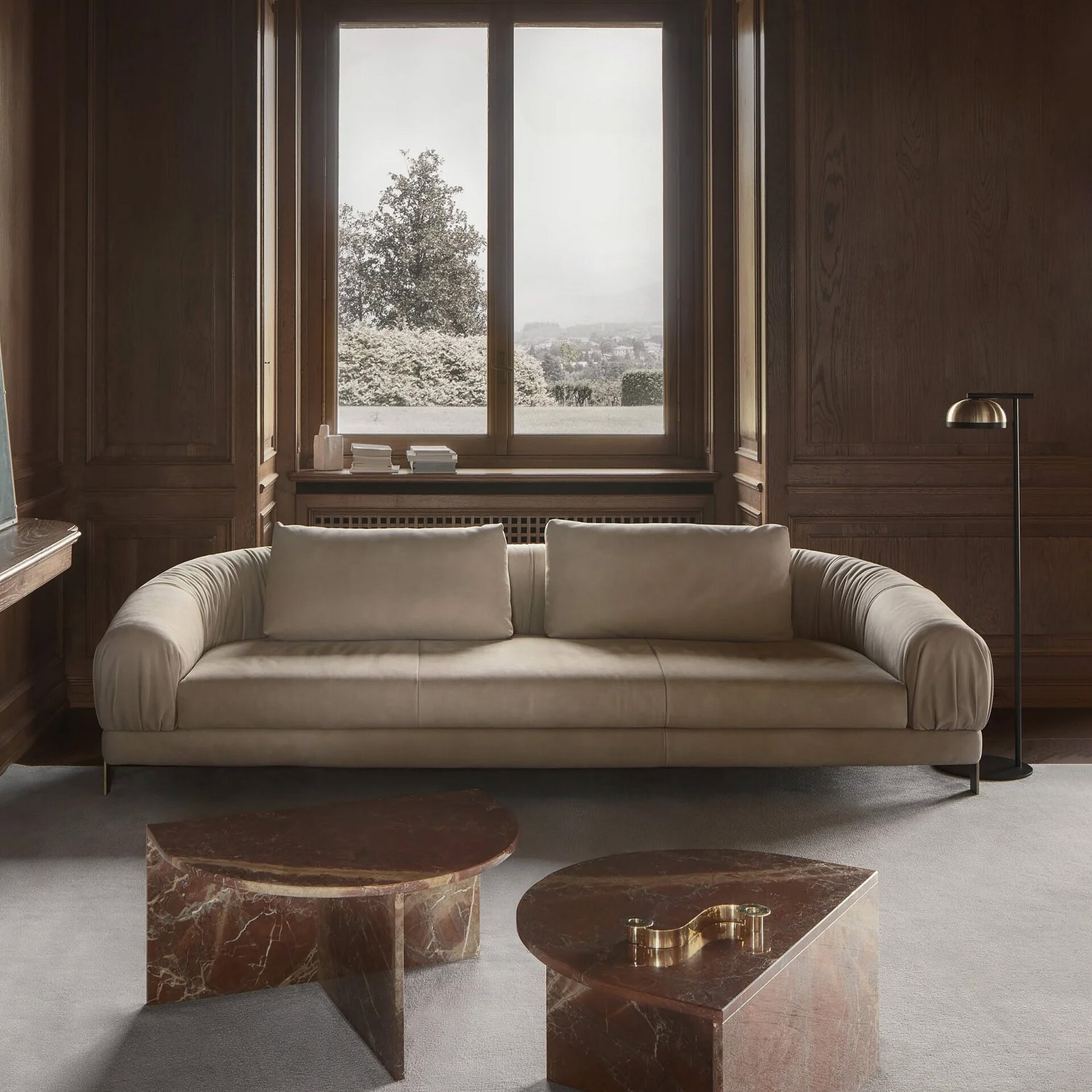 A luxurious living room with a light beige sofa, two reddish-brown marble coffee tables, dark wood paneling, and a large window overlooking a scenic landscape.