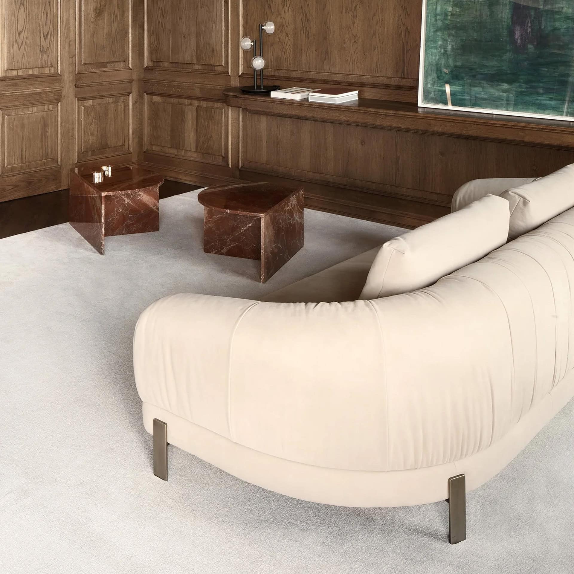 A luxurious living room featuring a cream curved sofa, two geometric red marble side tables, and dark wood paneled walls. A modern lamp, books, and a large green abstract painting are on a shelf above the tables.