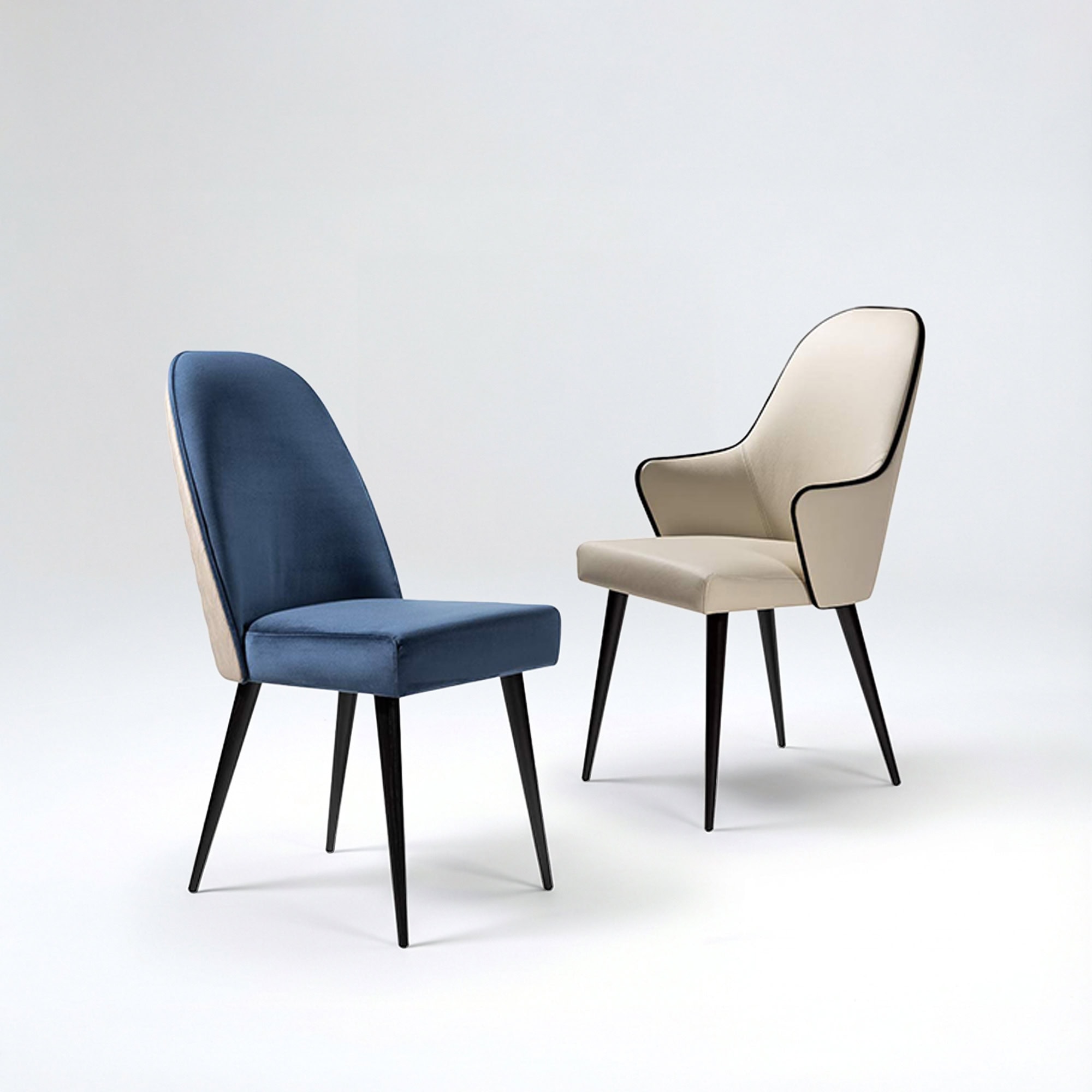 Two modern chairs on a white background: a blue velvet armless chair and a cream armchair with black piping, both with black tapered legs.